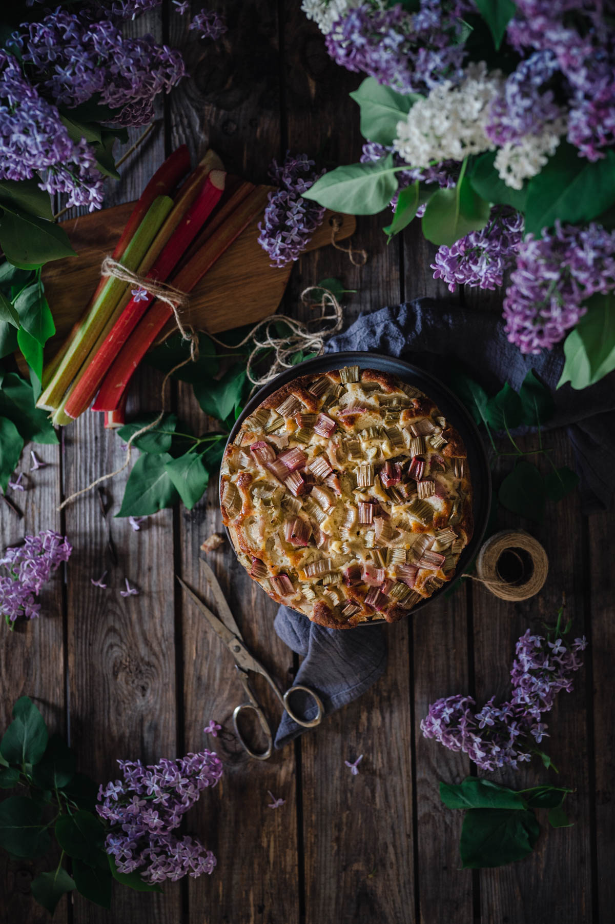 A flatlay of a rhubarb and ricotta cake on a wooden board. Lilacs and fresh rhubarb stalks.