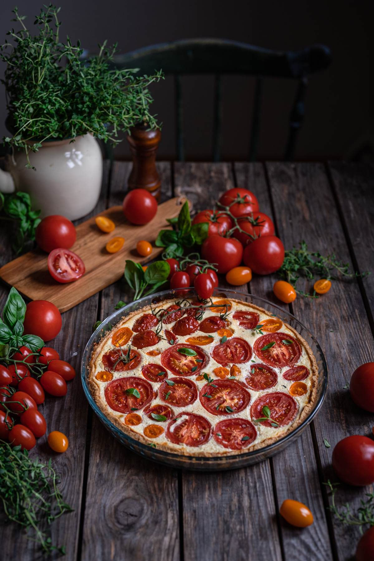 A whole tomato tart in a glassware tart pan surrounded by fresh tomatoes and herbs.