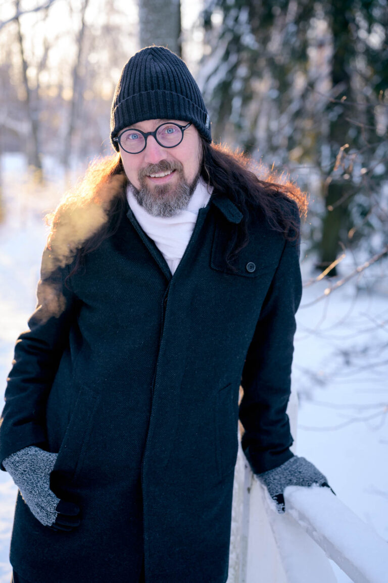 Mies nojaa lumiseen, valkoiseen aitaa. Miehen takaa paistaa aurinko matalalta. Miehellä on pitkät, kiharat hiukset, siniset silmät, mustat aurinkolasi. Mies hymyilee. Miehen päällä on musta pipo, musta takki, valkoinen kaulahuivi ja harmaat sormikkaat.